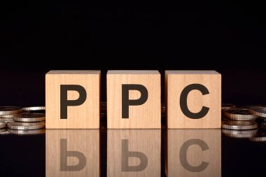PPC - Pay Per Click 'in kısaltması. Bozuk paralar, siyah arka plan ve iş konsepti içeren tahta küplerle ilgili yazı. Küplerdeki yazıtlar siyah tablonun yüzeyinden yansıyor. ön görünüm