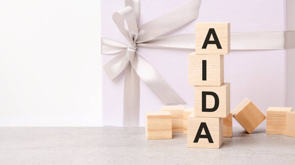 the AIDA text is laid out in a pyramid of wooden cubes. in the background is a paper gift box with a shiny white ribbon. AIDA - short for Attention Interest Desire Action