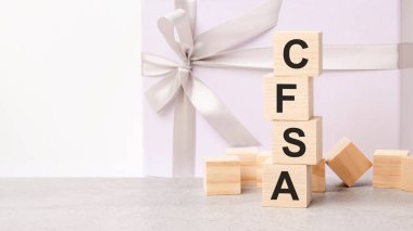 CFSA metni ahşap küplerden oluşan bir piramidin içinde. Arka planda parlak beyaz kurdeleli kağıt bir hediye kutusu var. CFSA - Sertifikalı Mali Hizmetler Denetçisinin kısaltması