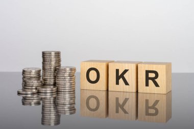 Tamam. Tahta küpler. Siyah arka planda bloklar var. Bozuk paralarla dolu desteler. Küplerdeki yazıtlar masanın yüzeyinden yansıyor. Seçici odaklanma. OKR - Hedefler ve Anahtar Sonuçların kısaltması