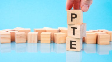 PTE metniyle ahşap küp bloğu tutan el. Küplerdeki yazılar yüzeyden yansıyor. Kopyalama alanı olan mavi arkaplan. PTE - Pearson İngilizce Testleri 'nin kısaltması