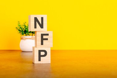 Üç tahta küp ve harfler. Sarı masada NFP, sağda yazı için yer var. Ön görüş kavramları, arka planda çiçekler. nfp - Çiftlik Dışı Maaş bordrosunun kısaltması