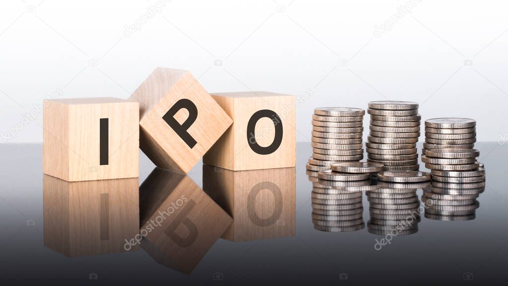 IPO - text is made up of letters on wooden cubes lying on a mirror surface, gray background. stacks with coins. inscription is reflected from the surface. IPO - short for Initial Public Offering
