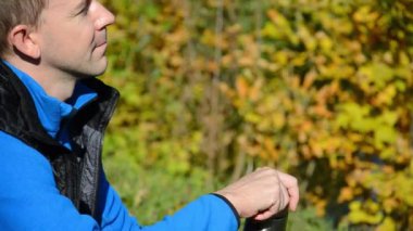 Beyaz adam sonbahar parkında oturup termostan çay içiyor. Sonbahar moda konsepti. Mavi ceketli erkek ve siyah jöle termosu açar ve kendini sarı ağaçların arka planında ılık içecekle ısıtır..