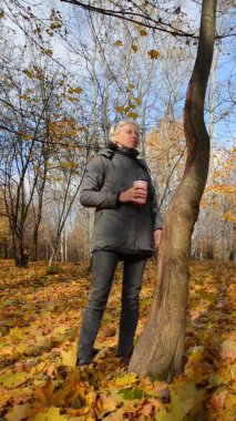 Güz parkında elinde bir fincan kahveyle duran güneş gözlüklü beyaz bir kadın. Sonbahar ormanında sarı yaprakların arasında bir kız. Rahatlama ve anın tadını çıkarma kavramı. Ruh hali dikey görüntüler.