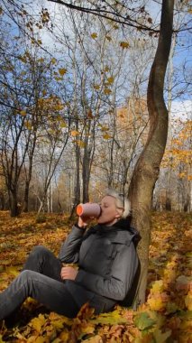 Beyaz bir kadın parkta oturuyor ve termostan sıcak su içiyor. Sonbahar ormanında sarı yaprakların arasında ağaca yaslanmış bir kız. Rahatlama ve anın keyfi kavramı.