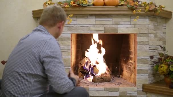 Un hombre con una camisa a cuadros se sienta solo junto a una chimenea en llamas — Vídeos de Stock