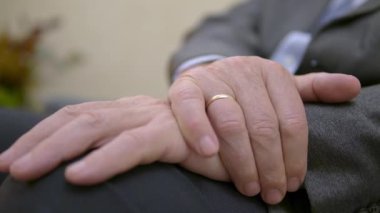 Takım elbiseli yetişkin bir adam dizinde katlanmış kollarıyla oturur.