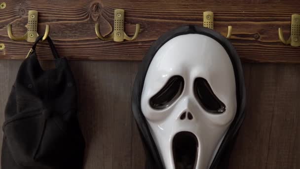 La horrible máscara de grito cuelga de un gancho junto con el tocado. Un maníaco con una máscara de miedo te está mirando. Una copia de la máscara de la película Scream. Halloween celebración — Vídeos de Stock