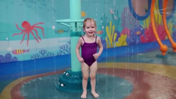 En liten flicka med blont hår i en lila baddräkt springer under vattenstrålarna av fontäner i vattenparken — Stockvideo