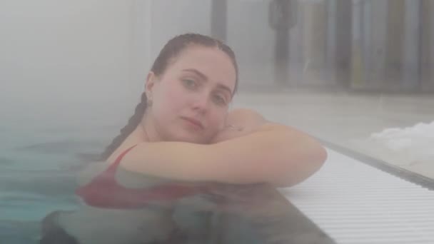Ένα νεαρό κορίτσι με σκούρα μαλλιά μαζεμένο σε μια πλεξούδα στέκεται στην πισίνα στο πλάι. Ζεστό εξωτερικό τζακούζι το χειμώνα και ατμός που βγαίνει από το νερό — Αρχείο Βίντεο