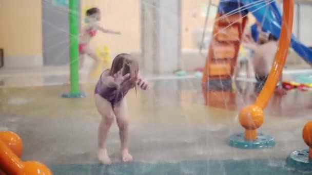 보라색 수영복을 입은 금발의 어린 소녀가 아쿠아 공원에 있는 분수 밑을 지나가고 있다 — 비디오