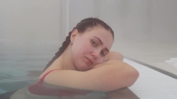 Uma jovem com cabelo escuro reunida em uma trança fica na piscina ao lado. jacuzzi ao ar livre quente em tempo de inverno e vapor saindo da água — Vídeo de Stock