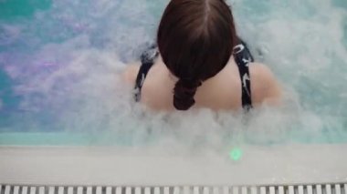 Koyu saçlı bir kız suda oturur ve su masajı sırtındaki suyun basıncına vurur.