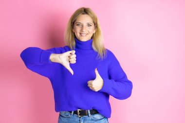 Genç ve güzel bir kadın pembe arka plan üzerine günlük süveter giyip baş parmak kaldırıyor, anlaşmazlık ve mutabakat ifadesi takınıyor. Çılgın çatışma