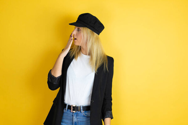 Beautiful woman wearing casual white t-shirt and a cap over isolated yellow background covering mouth with hand, shocked and afraid for mistake. surprised expression