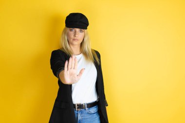 Güzel bir kadın, günlük beyaz tişört ve izole edilmiş sarı arka planda bir kep giyiyor ve avuç içiyle şarkı söylemeyi kesiyor..