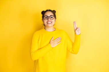 Genç, güzel bir kadın, izole edilmiş sarı arka planda rahat bir süveter giyiyor. Gülümsüyor, elleri göğsünde ve parmakları havada küfrediyor, sadakat yemini ediyor.