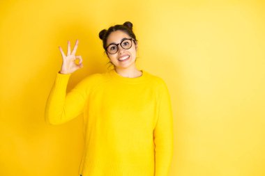 İzole edilmiş sarı arka plan üzerine sıradan süveter giyen genç güzel bir kadın tamam işareti yapıyor parmaklarıyla ve gülümsüyor, mükemmel bir sembol.