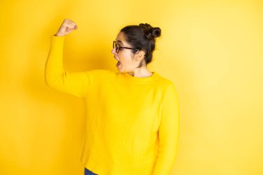 İzole edilmiş sarı arka planda sıradan bir süveter giyen güzel bir kadın gururla gülümseyen kol kaslarını gösteriyor.