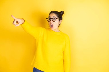 İzole edilmiş sarı arka plan üzerine sıradan süveter giyen genç ve güzel bir kadın parmağı önde, ağzı açık, şaşırmış, ön tarafta bir şey var.