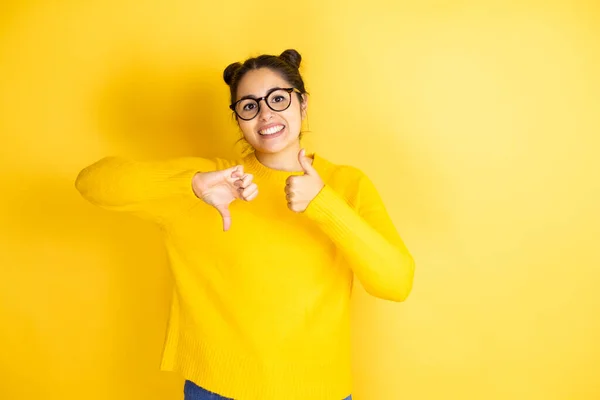 孤立した黄色の背景にカジュアルなセーターを着て若い美しい女性親指を上下に 意見の相違や合意表現を行います 狂気の葛藤 — ストック写真