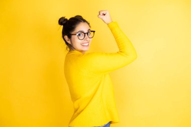 Genç, güzel bir kadın, izole edilmiş sarı arka plan üzerine günlük süveter giyiyor. Mutlu, tatmin olmuş ve güçlü hissediyor, kaslı ve formda. Spor salonundan sonra güçlü görünüyor.