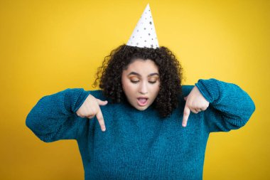 İzole edilmiş sarı arka plan üzerinde bir doğum günü şapkası takan genç ve güzel bir kadın şaşırmış, aşağı bakıyor ve parmaklarıyla işaret ediyor ve kollarını kaldırıyor.