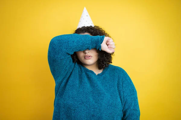 孤立した黄色の背景に誕生日の帽子をかぶっている若い美しい女性は 陽気で面白い腕で目を覆います ブラインドコンセプト — ストック写真
