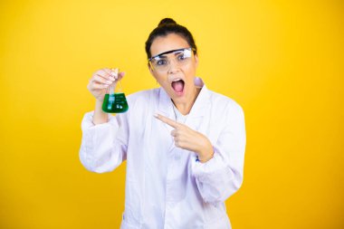 Bilim adamı üniforması giyen genç esmer kadın izole edilmiş sarı arka plan üzerinde test tüpü tutuyor, gülümsüyor ve test tüpünü işaret ediyor.