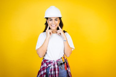 Kafkaslı genç bir kadın izole edilmiş sarı arka planda inşaatçı kıyafetleri giyiyor. Gülümsüyor ve parmaklarıyla ve ağzıyla işaret ediyor.