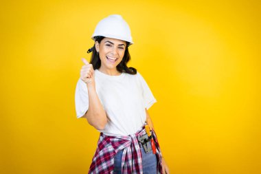 Kafkaslı genç bir kadın sarı arka plan başarısı tabelasının üzerine inşaat kıyafetleri takıyor. Elleriyle olumlu jest yapıyor, başparmağıyla gülümsüyor ve mutlu. Neşeli ifade