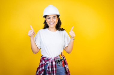 Kafkaslı genç bir kadın izole edilmiş sarı arka plan başarısı işareti üzerine inşaat kıyafetleri giyiyor. Elleriyle olumlu jest yapıyor. Gülümsüyor ve mutlu. Neşeli ifade