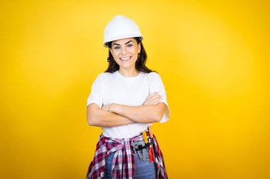 Kafkasyalı genç bir kadın kasketli ve inşaatçı kıyafetleri ile izole edilmiş sarı arka plan üzerinde mutlu bir yüz ile ayakta duruyor ve gülümsüyor kollarını kavuşturmuş dişlerini gösteren kendine güvenen bir gülümsemeyle.