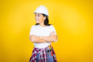 Kafkas şapkalı genç bir kadın, yalıtılmış sarı arka planda inşaatçı kıyafetleri giyiyor, rahat bir görünüm takınıyor, doğal bir yüz ifadesi ve kendine güvenen bir gülümsemeyle poz veriyor..