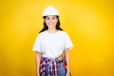 Kafkasyalı genç bir kadın, kasketli ve inşaatçı kıyafetleri ile izole edilmiş sarı arka plan üzerinde mutlu bir yüzle ayakta duruyor ve gülümsüyor dişlerini gösteren bir gülümsemeyle
