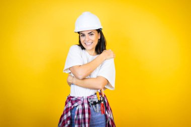 Kafkasyalı genç bir kadın kasketli ve inşaatçı kıyafetleriyle izole edilmiş sarı arka planda kendine sarılıyor mutlu ve pozitif, kendinden emin gülümsüyor.