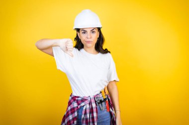 Kafkas şapkalı genç bir kadın ve inşaatçı kıyafetleri ile izole edilmiş sarı arka plan, kızgın yüz, negatif işaret baş parmağından aşağı hoşlanmadığını gösteriyor.