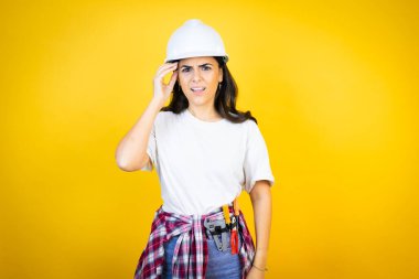 İzole edilmiş sarı arka plan kafa karışıklığı ve soru sorulmasına karşı kasketli ve inşaatçı giysileri giyen genç beyaz kadın. Şüpheyle karışık, kafa kafaya düşünerek.