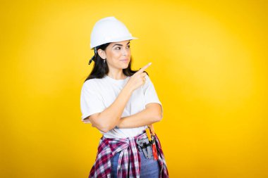 Kafkaslı genç bir kadın, sarı arka plan üzerinde inşaat kıyafetleri giyiyor. Kafası karışmış ve eli ve parmağı yana doğru uzanıyor.