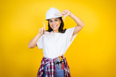 Kafkaslı genç bir kadın izole edilmiş sarı arka plan üzerinde inşaat kıyafetleri giyiyor ve gülümsüyor elleri ve parmakları mutlu yüzlü bir çerçeve yapıyor.