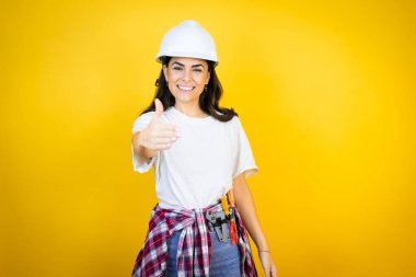 Kafkas şapkalı genç bir kadın izole edilmiş sarı arka planda inşaatçı kıyafetleri giyiyor. Dostça bir selamlama ve hoş geldin hediyesi olarak gülümseyerek el sıkışıyorlar.