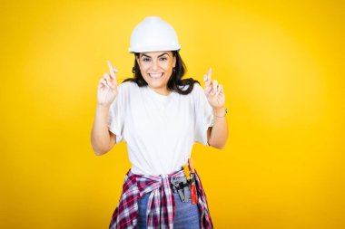 Beyaz şapkalı genç bir kadın, sarı arka plan üzerinde inşaatçı kıyafetleri giyiyor. Umut dolu bir gülümsemeyle işaret ediyor ve bakıyor.