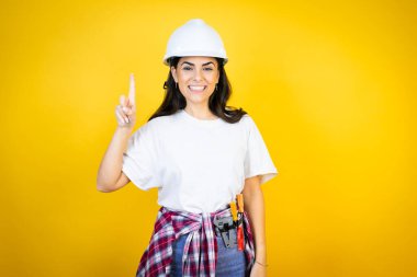 Kafkasyalı genç kadın kasketli ve inşaatçı kıyafetleri giyip izole edilmiş sarı arka planda bir numarayı işaret ederken kendinden emin ve mutlu bir şekilde gülümsüyor.
