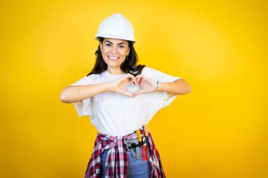 Kafkas şapkalı genç bir kadın izole edilmiş sarı arka planda inşaatçı kıyafetleri giyiyor. Aşkla gülümsüyor, kalp sembolü ve şekli elleriyle gösteriyor.