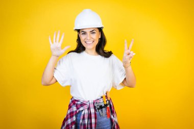 Kafkasyalı genç bir kadın kasketli ve inşaatçı kıyafetleri giyip izole edilmiş sarı arka plan üzerinde 7 numarayı işaret ederken kendinden emin ve mutlu bir şekilde gülümsüyor.
