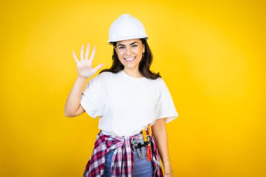 Kafkasyalı genç bir kadın kasketli ve inşaatçı kıyafetleri giyip izole edilmiş sarı arka planda beş numarayı işaret ederken kendinden emin ve mutlu bir şekilde gülümsüyor.