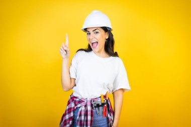 Kafkaslı genç bir kadın, sarı arka plan üzerinde inşaat kıyafetleri giyiyor. Parmağını kafasında gezdirip bir fikri olduğunu düşünüyor..