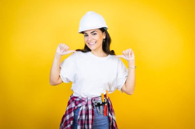 Kafkas şapkalı genç bir kadın ve sarı arka planda inşaatçı kıyafetleri giyiyor. Yüzünde bir gülümsemeyle, gururlu ve mutlu bir şekilde kendini işaret ediyor..