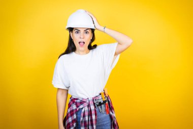 Kafkaslı genç bir kadın, sarı arka plan üzerinde inşaat kıyafetleri giyiyor. Bir elini başının üstüne koyup sanki bir şey unutmuş gibi gülümsüyor.